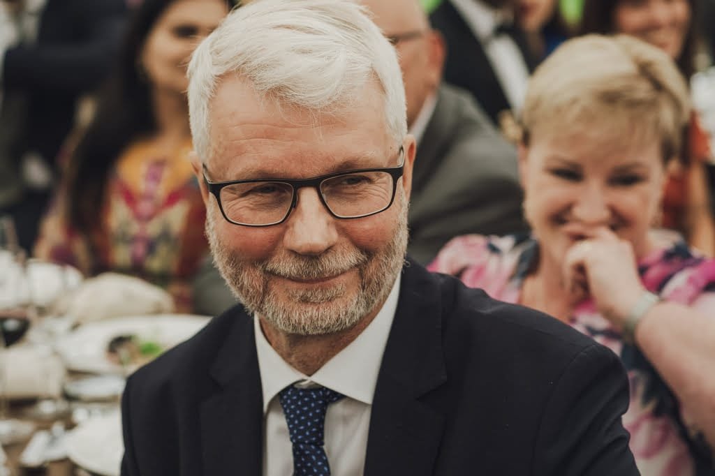 A man with white hair, glasses, and a beard, wearing a dark suit and blue tie, is smiling at an event held at Farleigh House Wedding Photographer venue, with people seated around him.