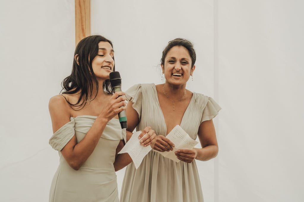 Two women in formal dresses stand close together, both holding notes. The woman on the left is speaking into a microphone and smiling. They appear to be making a speech, beautifully captured by the Farleigh House Wedding Photographer.