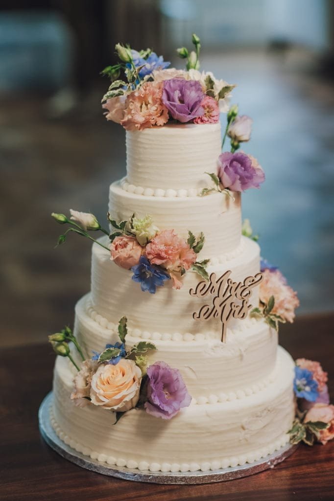 A four-tiered white wedding cake decorated with various flowers, including purple and pink blossoms, stands elegantly on a wooden table. Captured beautifully by the Farleigh House Wedding Photographer, the cake topper proudly reads "Mr & Mrs.