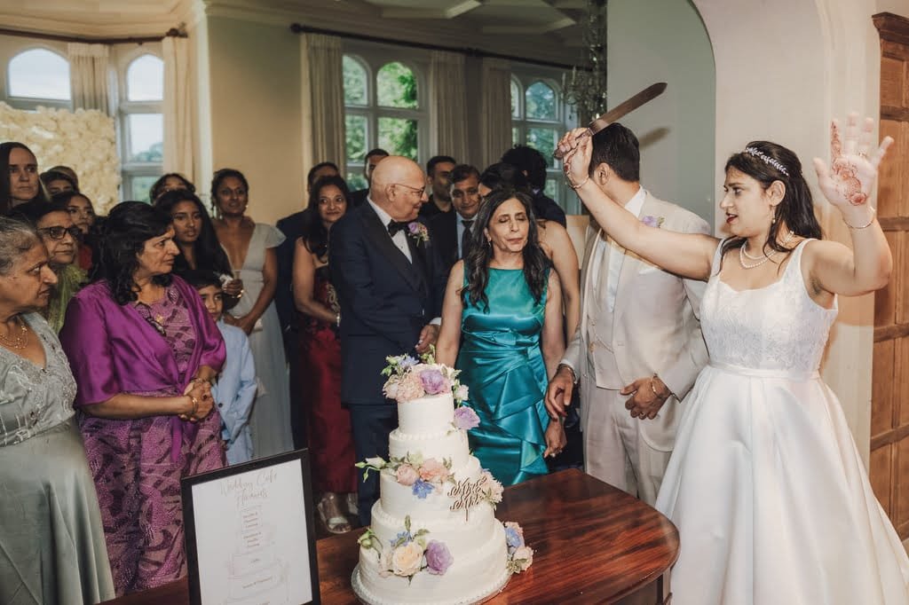 A bride in a white dress holds a knife near a decorated wedding cake, preparing to cut it. Guests in formal attire stand around her, watching and smiling, as the Farleigh House Wedding Photographer captures every precious moment.