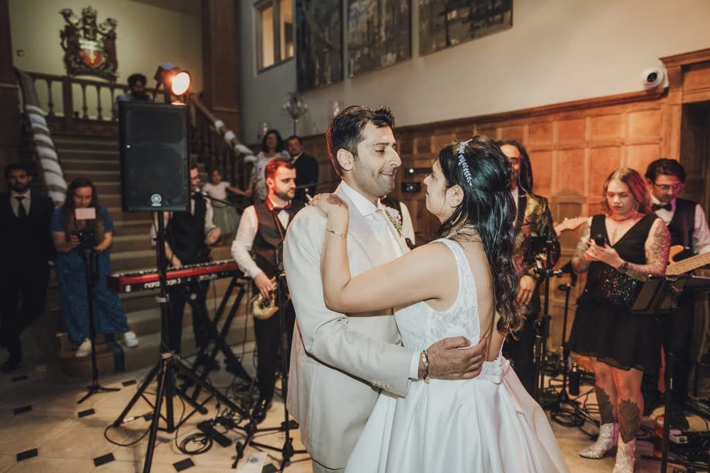 A couple in formal attire is dancing at their wedding reception, captured beautifully by a Farleigh House Wedding Photographer. A band with various instruments plays energetically on stage, while guests watch from the nearby staircase and the floor.