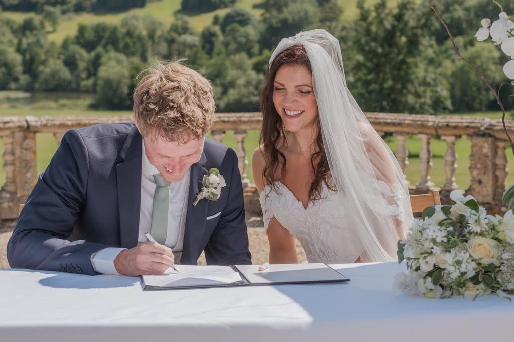 A groom signs a document while a bride in a white dress and veil smiles next to him. They are outdoors on a sunny day with greenery, the elegant stone balustrade of Orchardleigh House in the background.