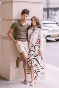 A man and woman posing together on a city sidewalk for their complimentary engagement photo shoot, both smiling and dressed in casual summer attire.
