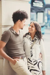 A smiling couple leaning against a wall during their complimentary engagement photo shoot, looking at each other with affection.