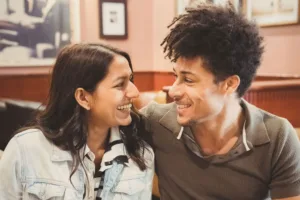 Two people smiling at each other in a cozy setting during their complimentary engagement photo shoot.