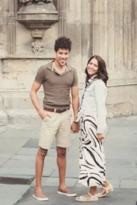 A smiling couple standing together on a stone-paved street, with historic architecture in the background during their complimentary engagement photo shoot.