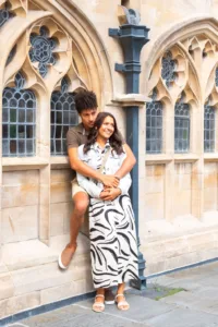 A couple posing with a hug against a historical building's wall with gothic windows during their complimentary engagement photo shoot.