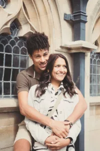 A smiling couple embracing in front of an arched stone window during their complimentary engagement photo shoot.