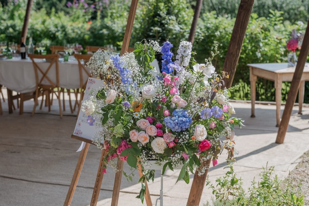 A large floral arrangement with various colored flowers including roses, hydrangeas, and baby's breath is displayed under a tented area at PYTHOUSE WEDDINGS, with tables and chairs in the background.