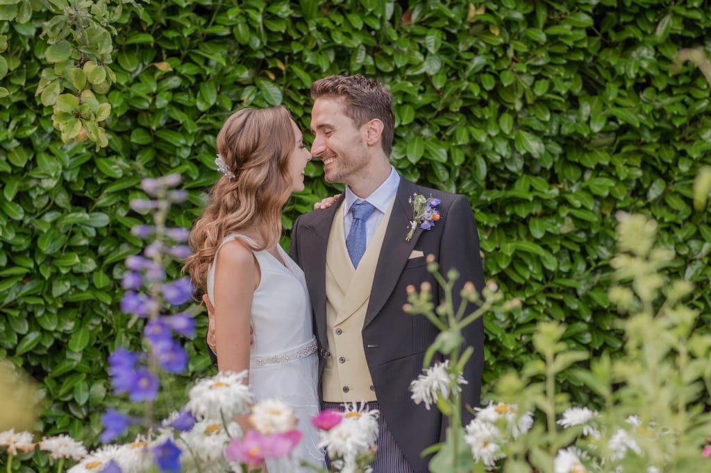 A couple, dressed in wedding attire, stands closely facing each other in front of a leafy green background at PYTHOUSE WEDDINGS, surrounded by colorful flowers.