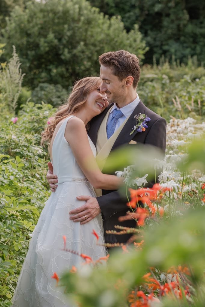A bride and groom stand closely together, smiling amidst a garden with greenery and colorful flowers at Pythouse Weddings. The bride wears a white dress, and the groom is in a navy suit with a yellow vest and blue tie.