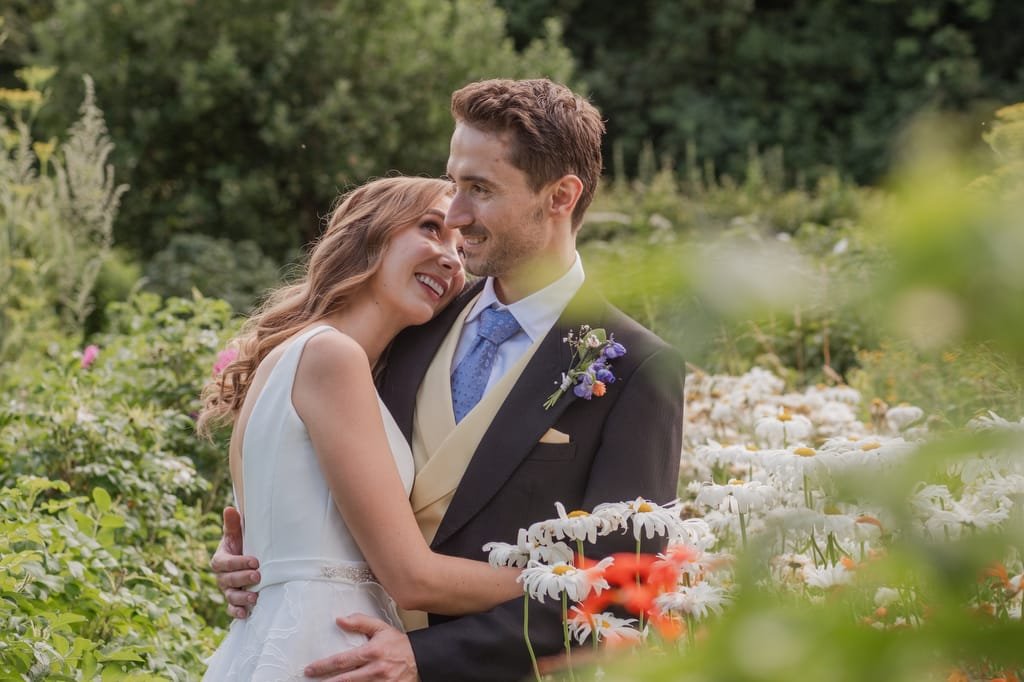 A couple dressed in wedding attire embraces in a garden filled with daisies and greenery at PYTHOUSE WEDDINGS.