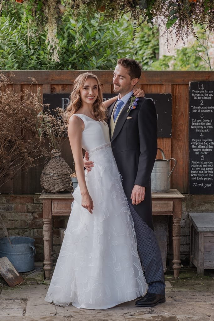 A couple stands together, smiling, in front of a rustic background with greenery and wooden elements at Pythouse Weddings. The woman wears a white dress, and the man wears a suit with a floral boutonniere.