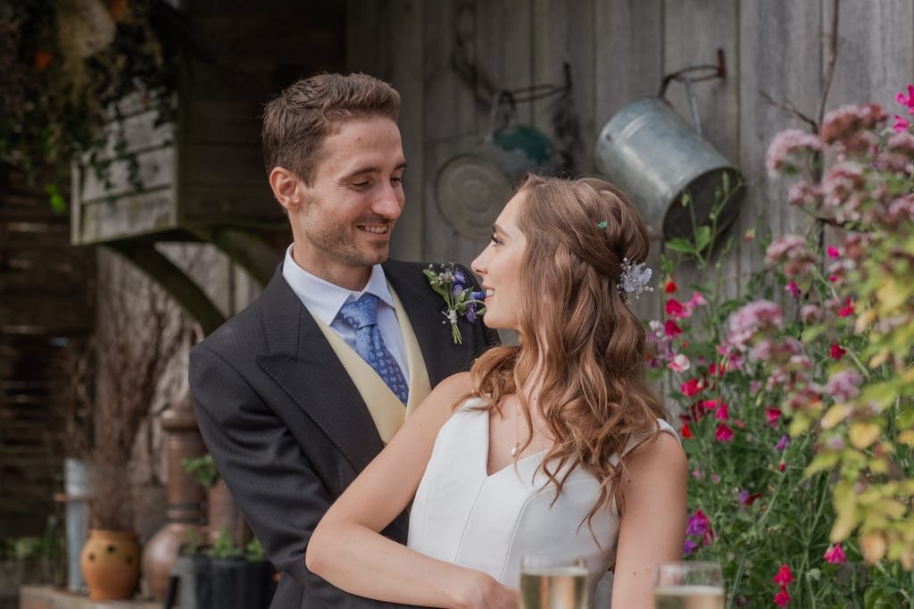 A couple, dressed formally, stands outdoors in front of a rustic wooden structure adorned with flowers and garden tools, looking at each other and smiling. Glasses of champagne are in the foreground, capturing the romantic essence of PYTHOUSE WEDDINGS.