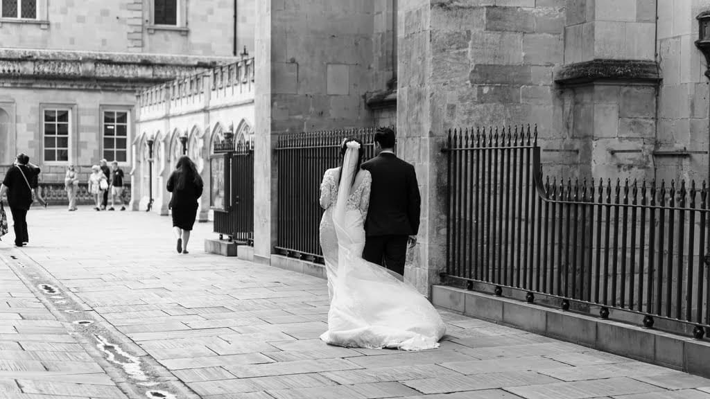 A bride and groom walk together down a cobblestone pathway beside the historic Guildhall Bath, a stone building. Other people are seen in the background. The image is in black and white.