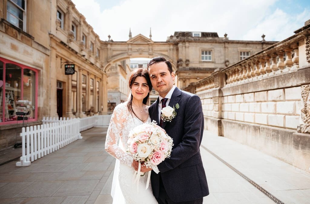 A bride in a white lace dress and groom in a dark suit pose together with a bouquet in front of the historic Guildhall Bath on a sunny day.