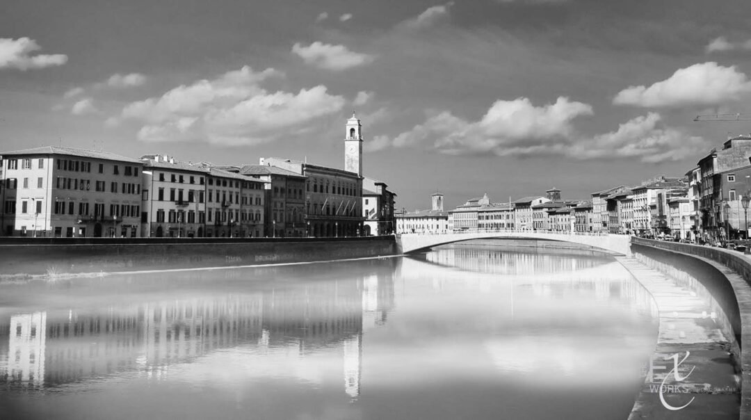 A black and white photo captures buildings and a clock tower lining a river, with a bridge crossing over the water under a partly cloudy sky—an elegant showcase of Black & White Film Photography.