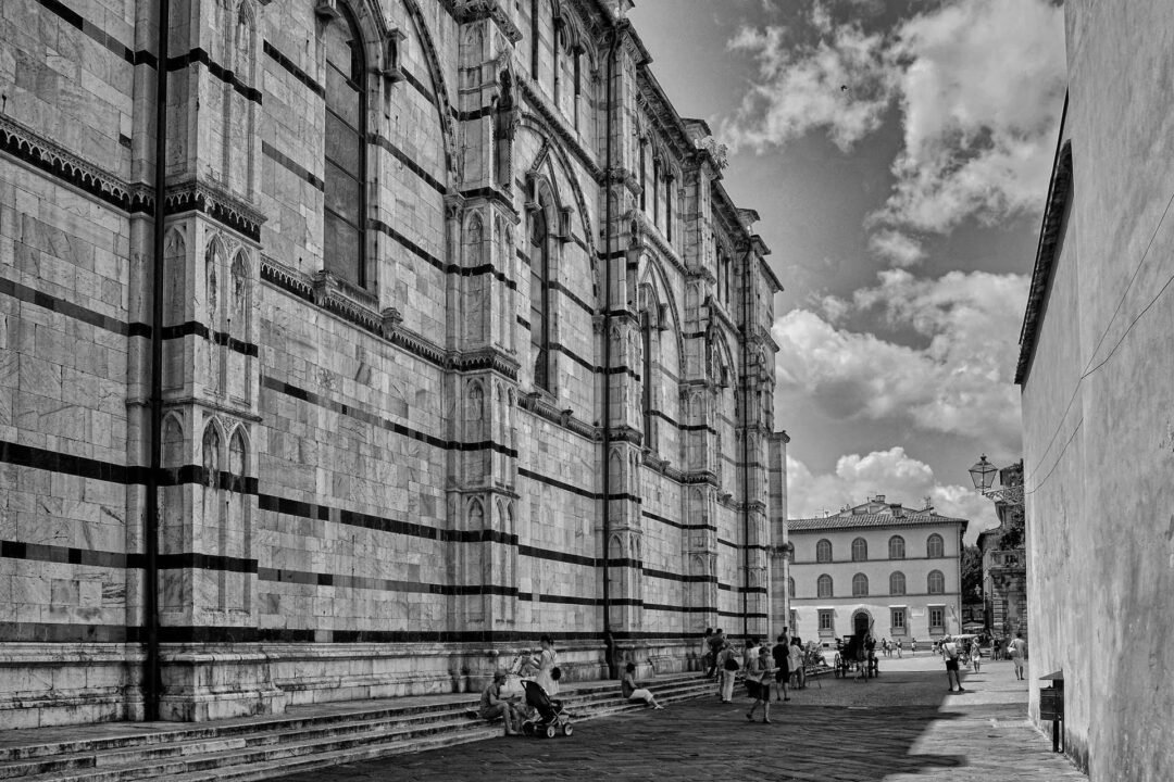 People walk and sit near the large stone facade of a historic building with gothic arches on a sunny day; another building is visible in the background. The scene is beautifully captured in classic Black & White Film Photography.