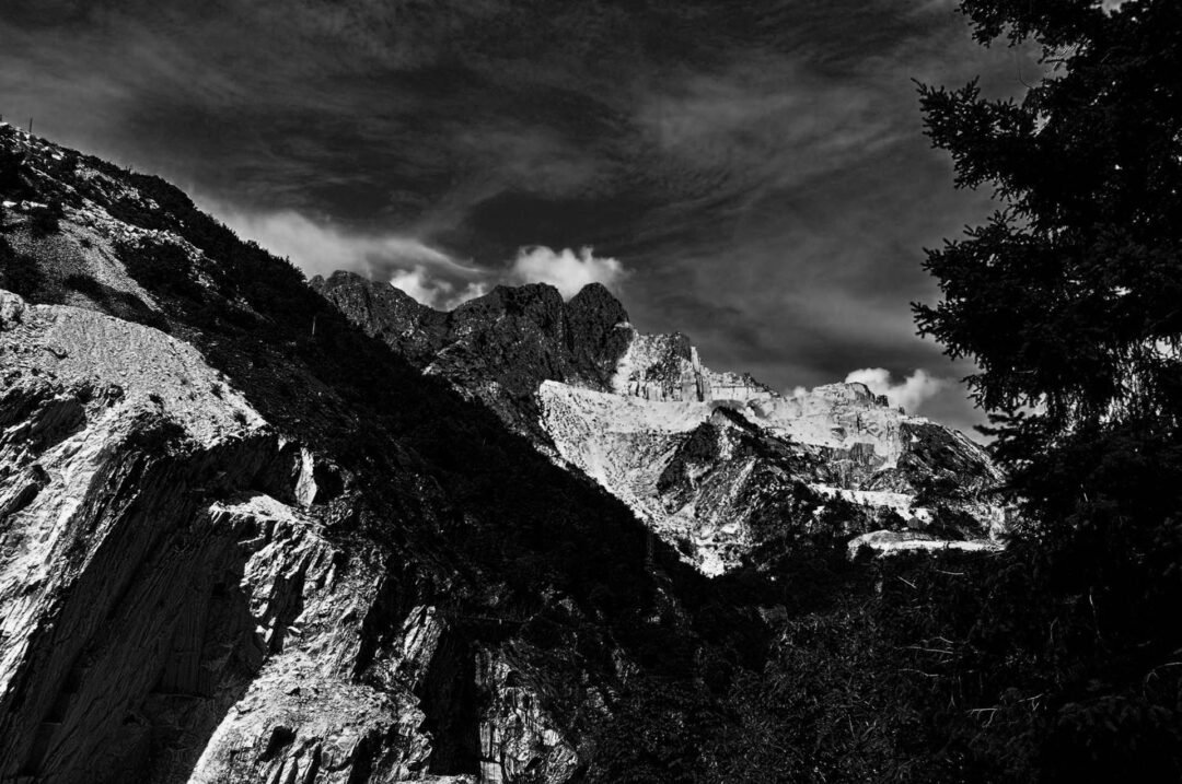 Black and white film photography captures rugged mountain peaks beneath a dramatic sky, with trees artfully framing the right side of the image.