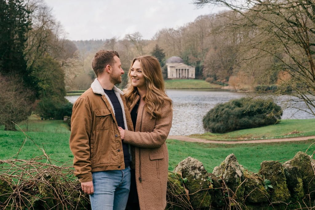 A portrait photography moment captures a couple standing outdoors in front of a stone wall with a serene lake and temple-like structure in the background. They are facing each other, smiling, with tree-covered hills surrounding them.