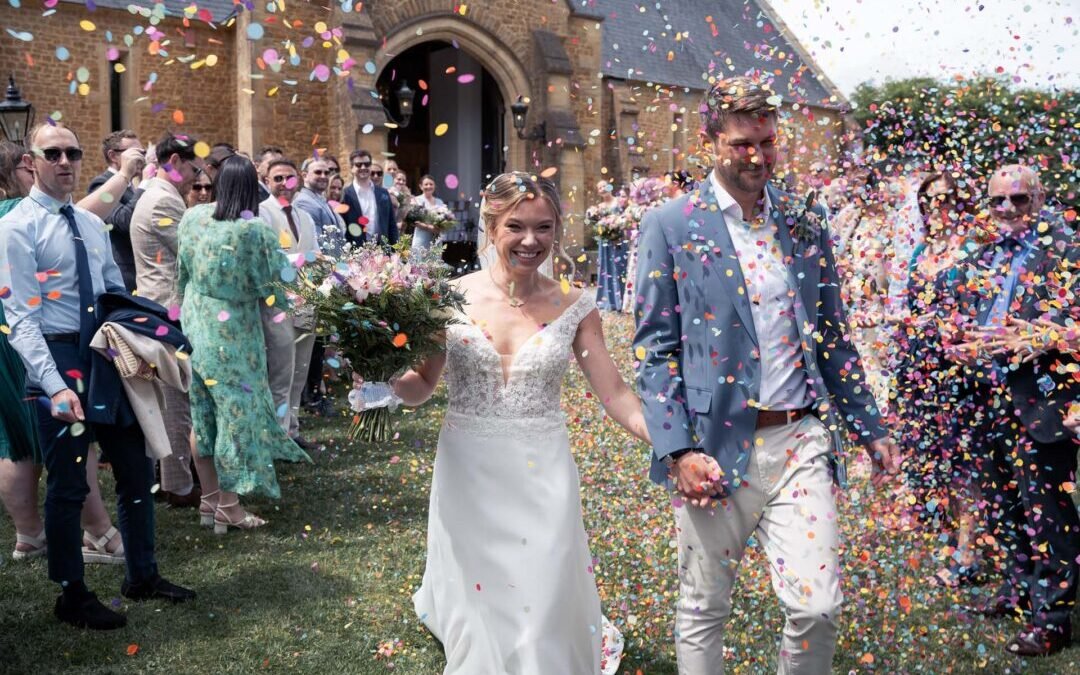 A bride and groom walk outside a church as guests throw colorful confetti; both are smiling and holding hands, surrounded by well-dressed attendees—capturing the joy of Haselbury Mill Weddings.