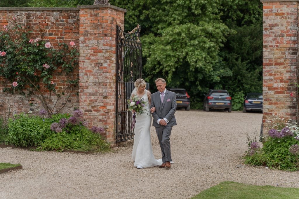 A bride in a white dress and veil walks arm in arm with a man in a gray suit and pink tie through a garden gate with brick pillars, heading towards the camera, beautifully captured by the weddings photographer at Elmhay Park.