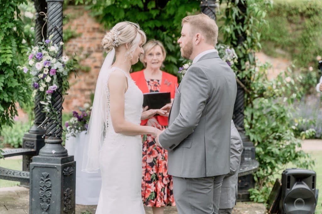 A bride and groom stand facing each other, holding hands, under an ornate gazebo adorned with flowers. As an officiant in a floral dress and red jacket conducts their wedding ceremony, the Weddings Photographer at Elmhay Park captures every cherished moment.