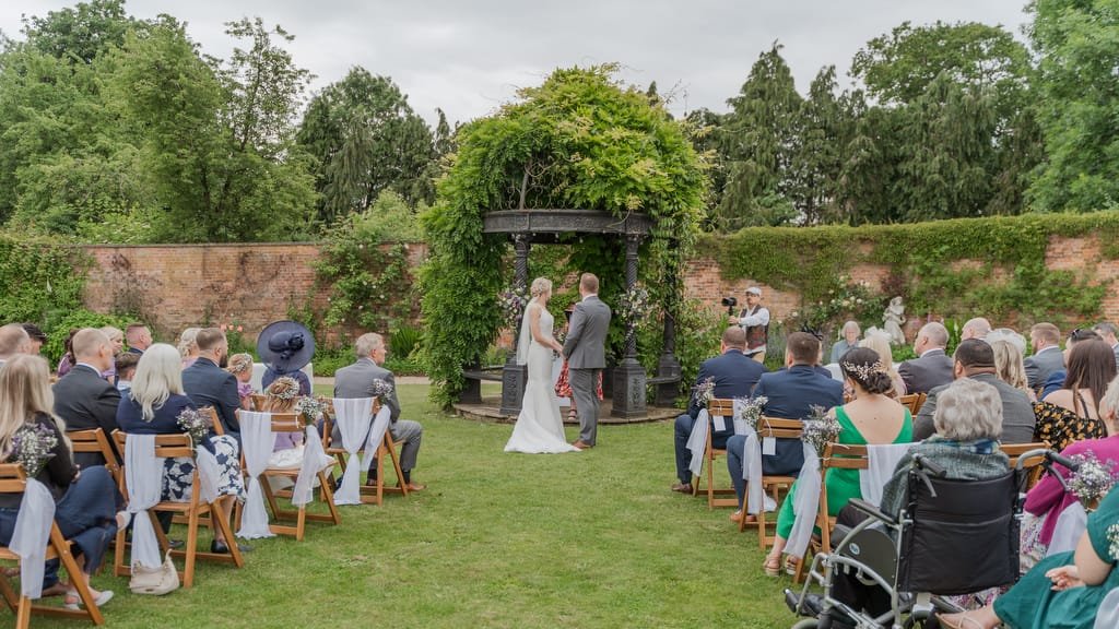 A couple stands under a gazebo with an officiant, exchanging vows in front of seated guests at an outdoor wedding ceremony. The skilled weddings photographer captures the moment amidst the serene backdrop of trees and a brick wall covered in greenery at Elmhay Park.