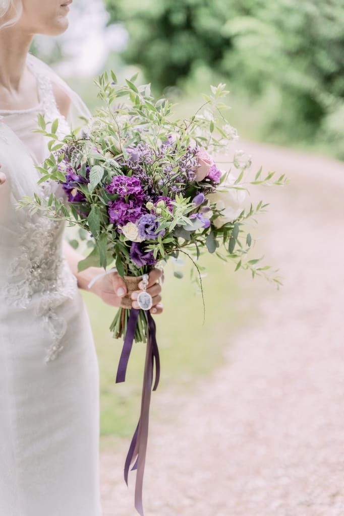 A bride in a white dress holds a bouquet of purple, pink, and green flowers with a winding path and lush greenery in the background, perfectly captured by a talented weddings photographer at Elmhay Park.