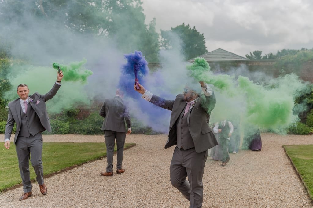Men in suits holding smoke bombs emit green and blue smoke in a garden setting at Elmhay Park, with others in the background partially obscured by the colorful clouds. This whimsical scene is captured beautifully by a skilled weddings photographer.
