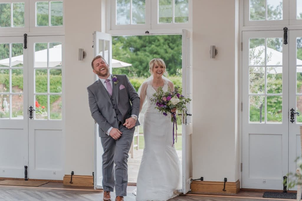 A bride and groom are entering a room through double doors, smiling and holding hands. The bride holds a bouquet of purple and white flowers. Capturing this enchanting moment, the weddings photographer at Elmhay Park ensures every detail is preserved with elegance.