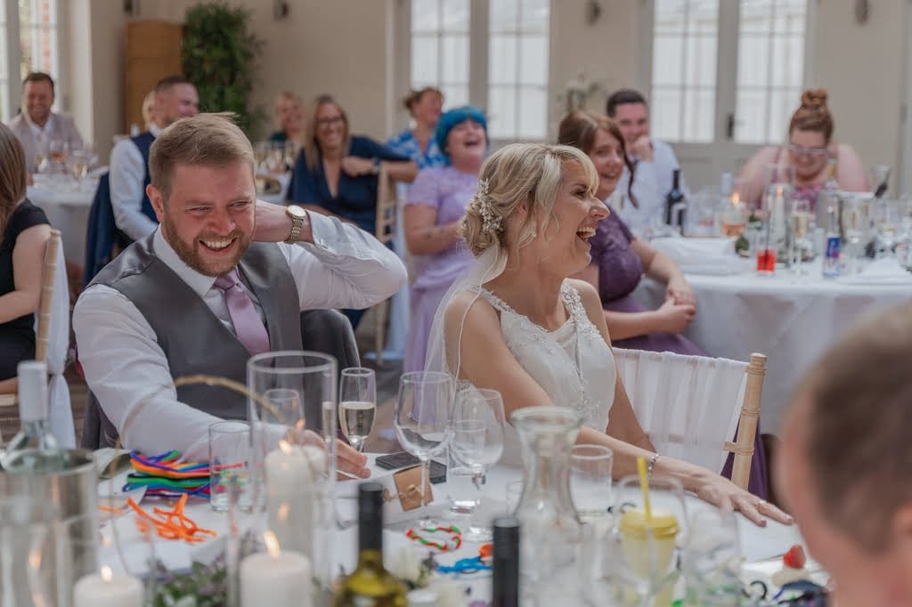 A bride and groom are seated at a table, smiling and laughing with their guests at Elmhay Park during a charming wedding reception. People in the background, captured beautifully by the weddings photographer, are also enjoying the moment.