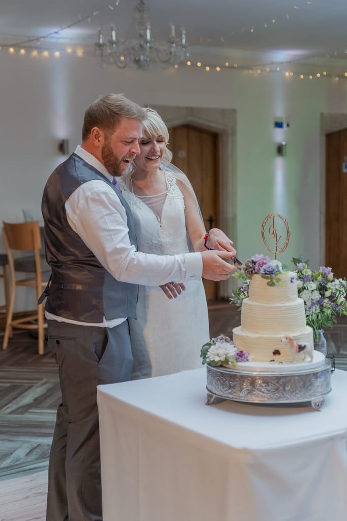 A man and woman in wedding attire smile as they cut a floral-decorated, tiered wedding cake on a round table, perfectly captured by the weddings photographer at Elmhay Park.