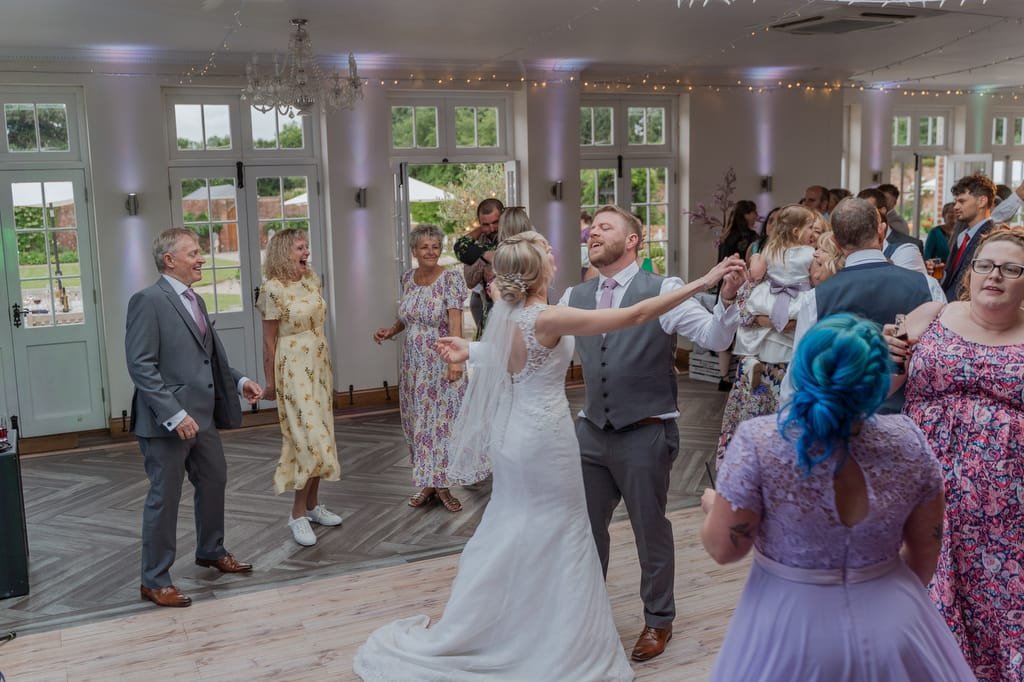 A bride and groom dance surrounded by guests in a bright reception hall, with some people clapping and others chatting. The setting, beautifully captured by the weddings photographer at Elmhay Park, includes large windows, string lights, and floral decorations.