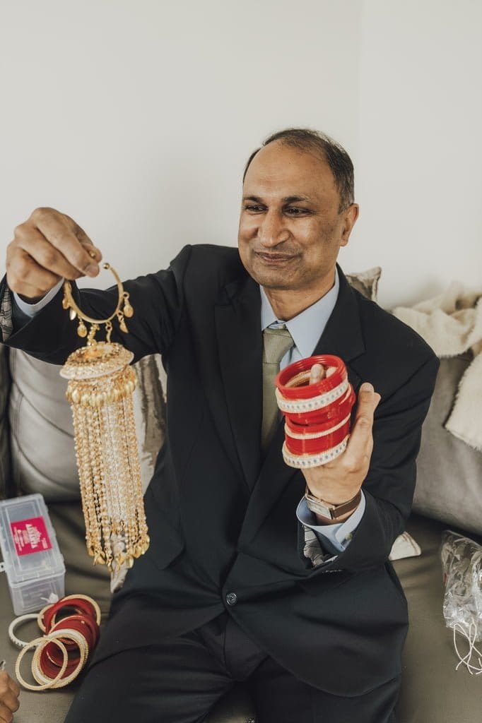 A man in a suit is sitting on a couch, holding up red bangles in one hand and a set of gold-colored chandelier earrings in the other, smiling—showcasing wedding traditions from around the world.