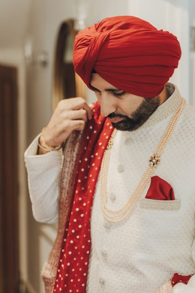 A man dressed in traditional attire with a red turban and a white sherwani adjusts his red and gold patterned shawl, embodying wedding traditions from around the world.