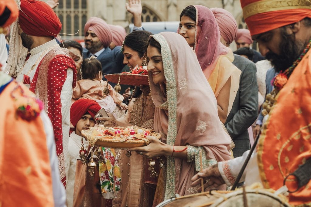 A group of people dressed in traditional attire participates in a cultural ceremony, reflecting wedding traditions from around the world, with one woman holding a decorated plate and others wearing turbans and colorful garments.