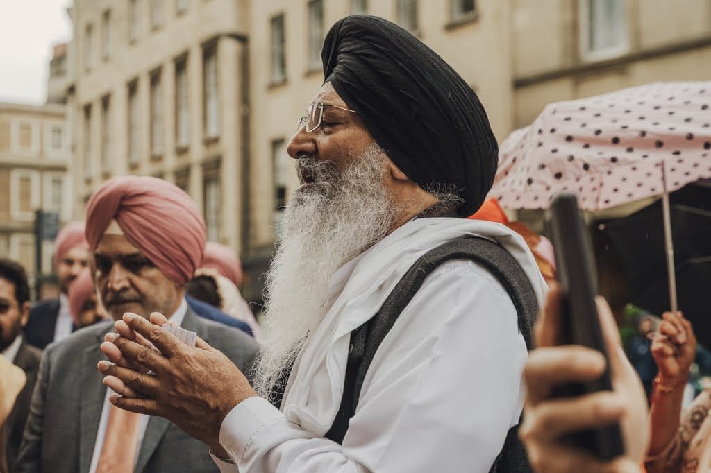 A man with a black turban and white beard stands among a crowd, clapping. Beside him is another man in a pink turban. As part of wedding traditions from around the world, people are holding umbrellas, adding to the festivity, while a smartphone is visible in the foreground capturing the moment.