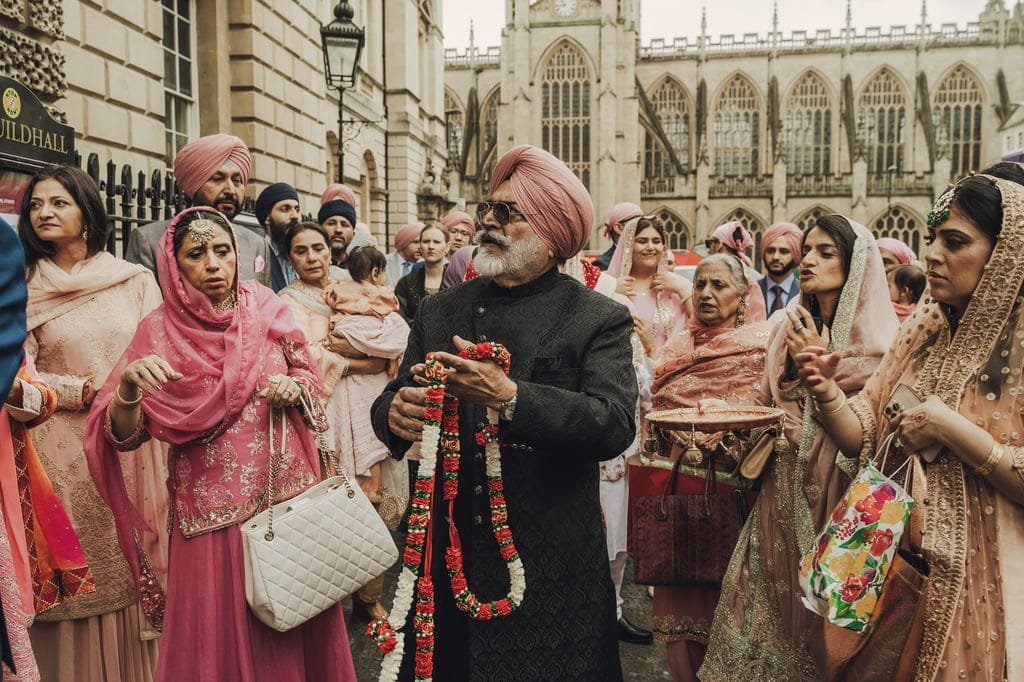 A man in traditional attire holds a flower garland, surrounded by a group of people in colorful clothing, in front of a historic building. Everyone looks engaged in an outdoor ceremony, celebrating wedding traditions from around the world.