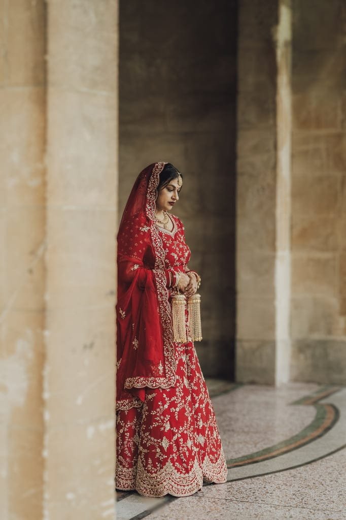 A woman in a red and gold traditional outfit and veil stands next to a column in a stone-arched walkway, holding decorative tassels, embodying the elegance of wedding traditions from around the world.