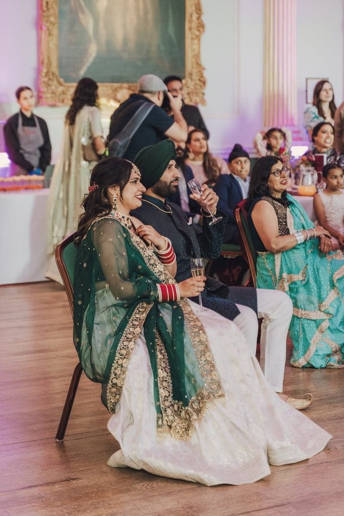 A seated couple in traditional attire watches an event, with other attendees in the background, celebrating wedding traditions from around the world at a beautifully decorated venue.