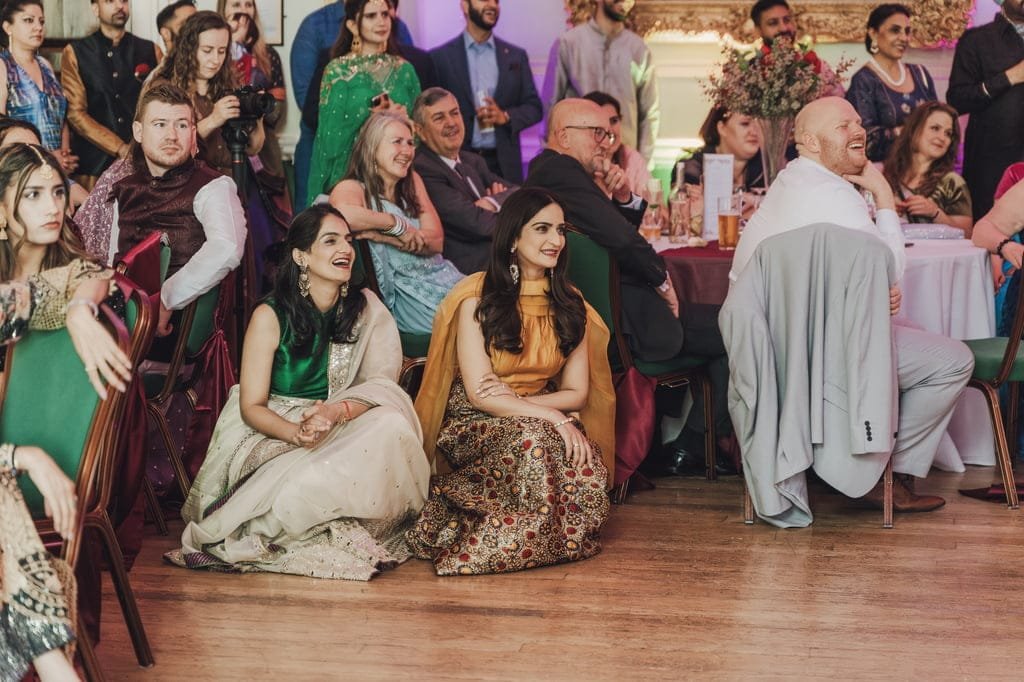 A group of people, including two women seated on the floor in traditional attire, watch an event with expressions of amusement and interest, showcasing wedding traditions from around the world.