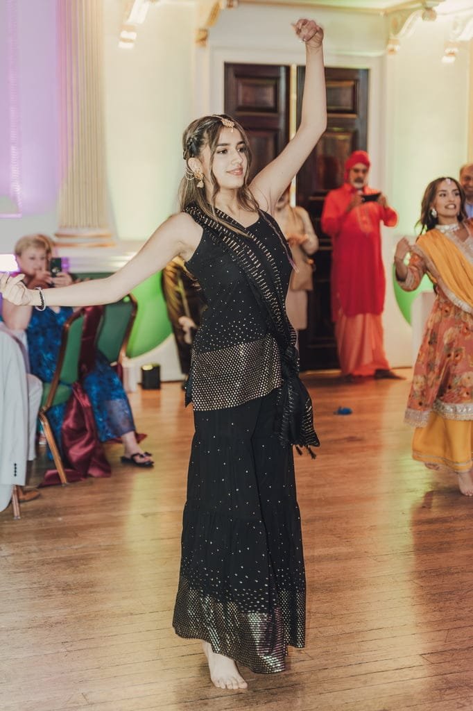 A woman in a black outfit dances barefoot in an indoor setting, embodying wedding traditions from around the world, while guests look on.