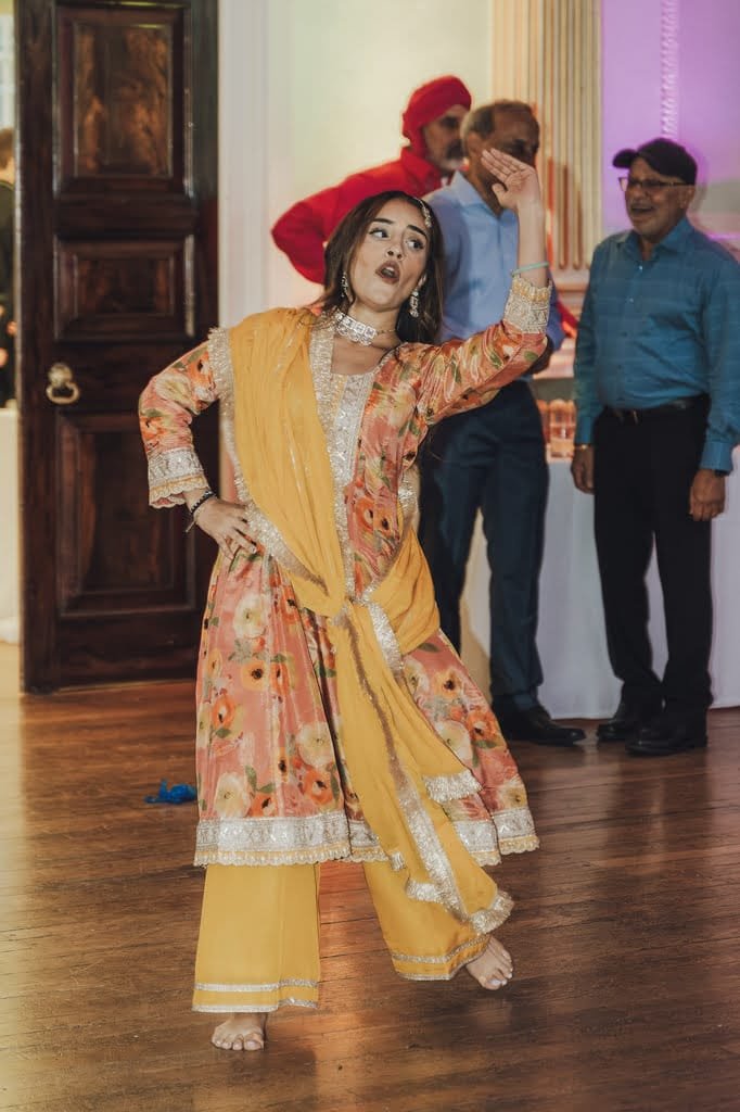 A woman in a vibrant traditional outfit performs a dance, raising one arm. Three men, one in a red turban and two in blue shirts, watch in the background, showcasing wedding traditions from around the world.