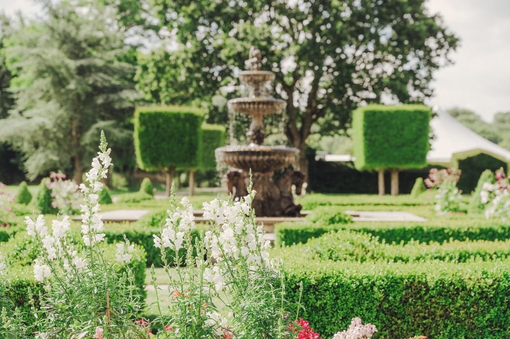 A formal garden featuring neatly trimmed hedges, blooming flowers, and a central tiered fountain with trees in the background—an idyllic setting perfect for a Farleigh House wedding photographer to capture unforgettable moments.