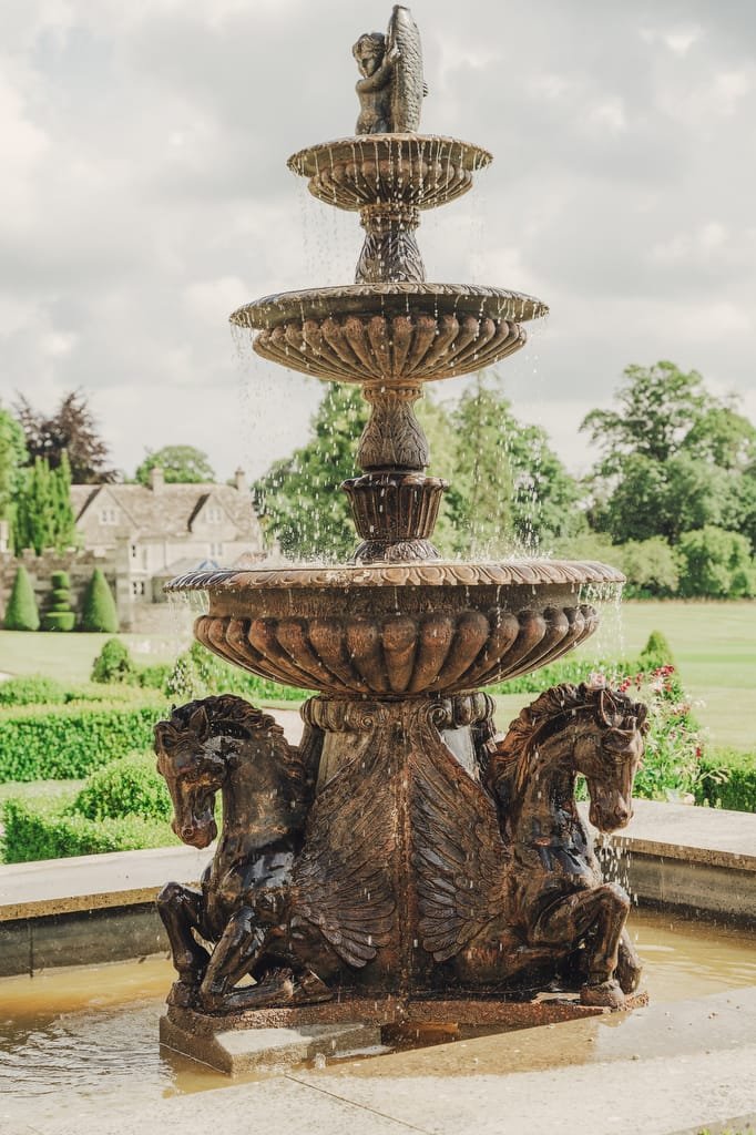 A three-tiered stone fountain with water flowing is adorned with sculptures of four horses at its base, set against a backdrop of trees and Farleigh House, making it an idyllic location for a wedding photographer.