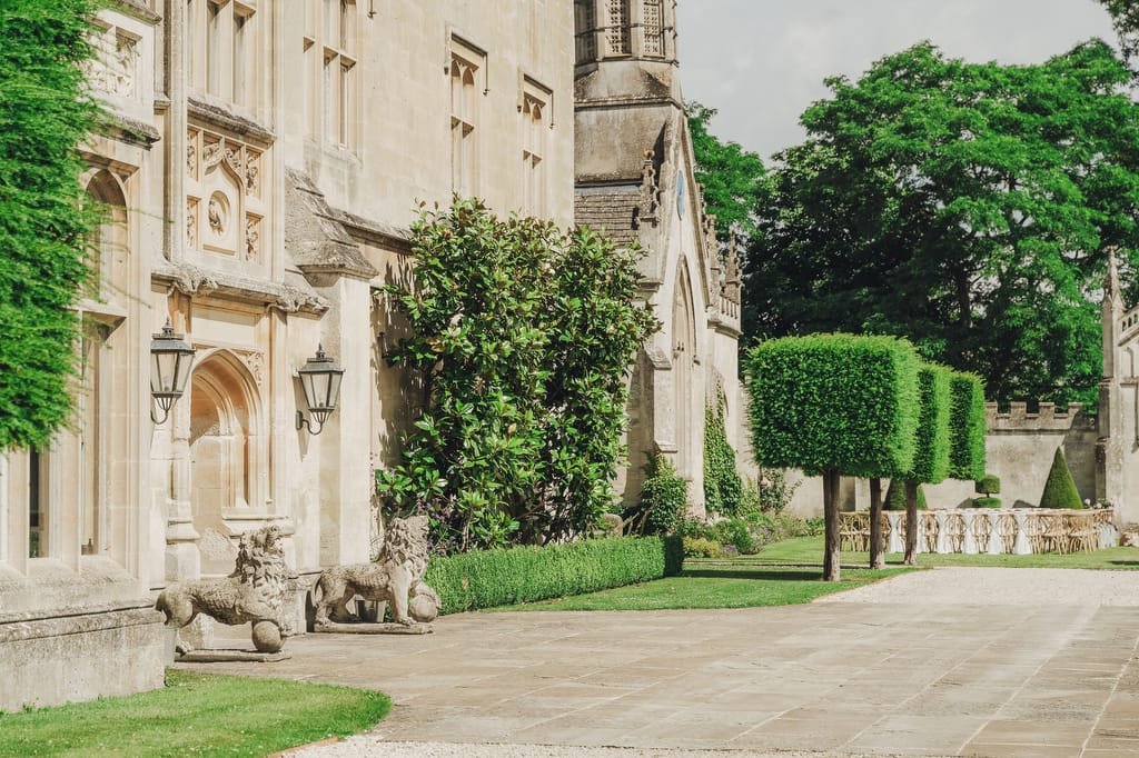 A historic stone building with ornate details, topiary trees, and lion statues near the entrance. The scene includes neatly trimmed hedges and trees, with a paved pathway leading along the side—an idyllic setting for any Farleigh House Wedding Photographer to capture timeless moments.
