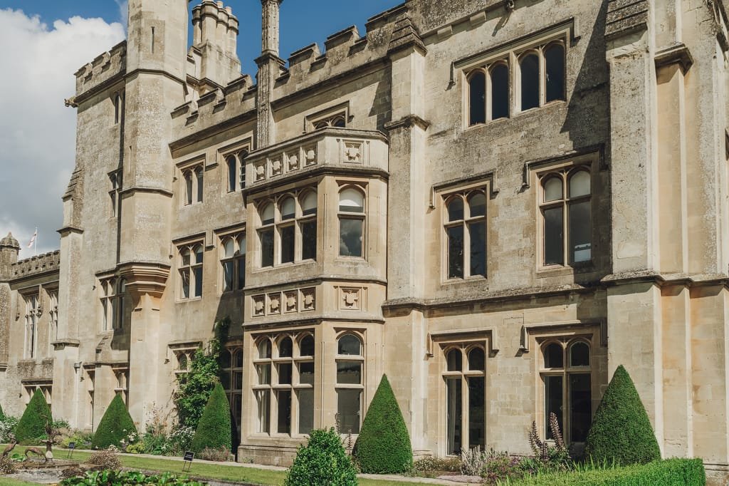 A historic stone building with multiple tall windows and turrets, Farleigh House stands elegantly surrounded by neatly trimmed hedges and a manicured garden under a partly cloudy sky. It's the perfect backdrop for any wedding photographer seeking timeless beauty.