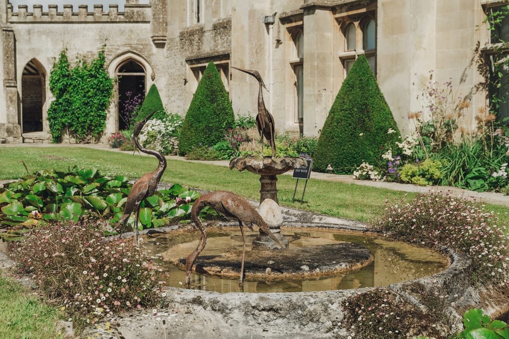 A small stone fountain with bronze bird statues is surrounded by greenery, flowers, and trimmed pyramid-shaped bushes in the garden of a historic stone building, creating a picturesque setting perfect for a Farleigh House Wedding Photographer.