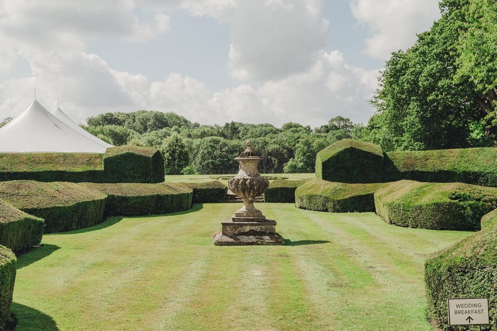 A formal garden with manicured hedges surrounds an ornate stone urn. A large white tent is visible in the background. A sign reading 